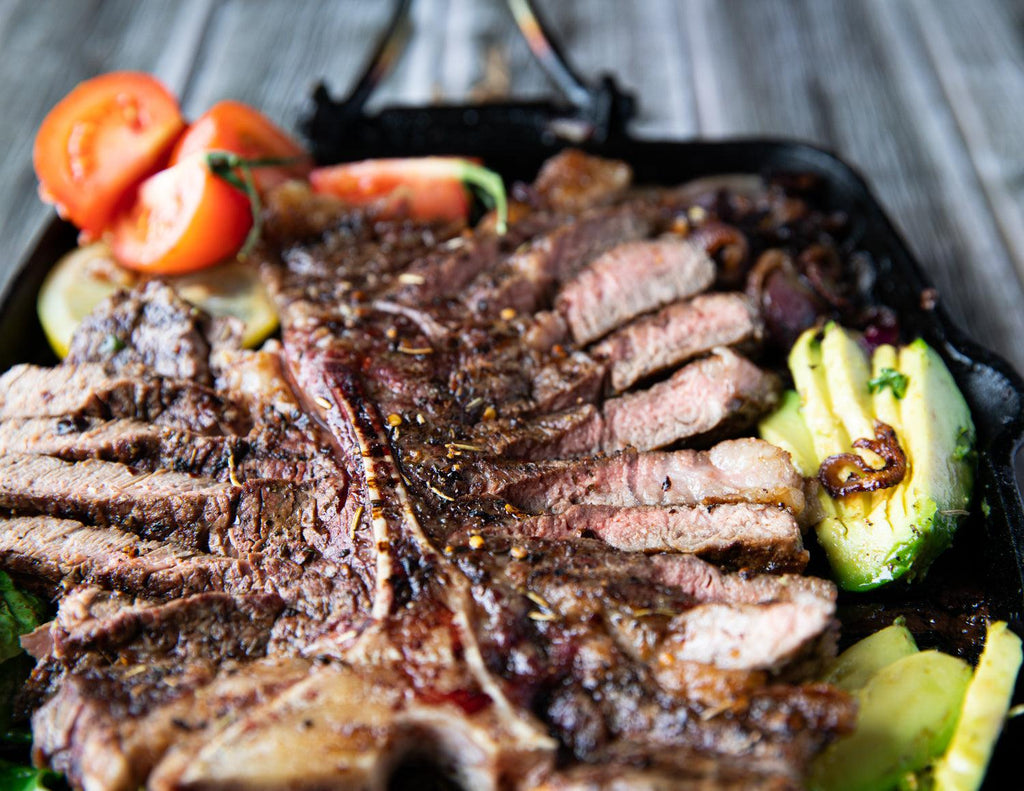 A Porterhouse Steak in a cast iron skillet with avocados, tomatoes and sauteed onions.